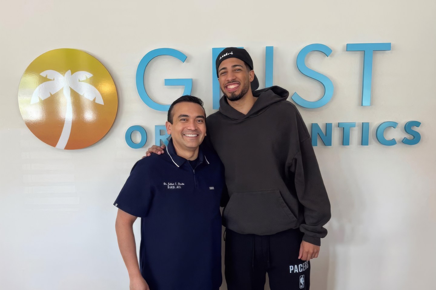 Smiling orthodontist and patient posing together in front of Geist Orthodontics logo, showcasing a welcoming atmosphere and community engagement.
