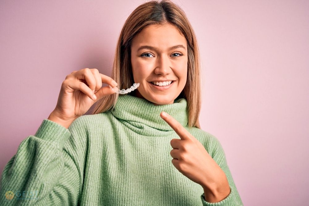 Smiling young woman in a green sweater pointing at a clear aligner in her hand after understanding does insurance cover Invisalign in Indianapolis, IN