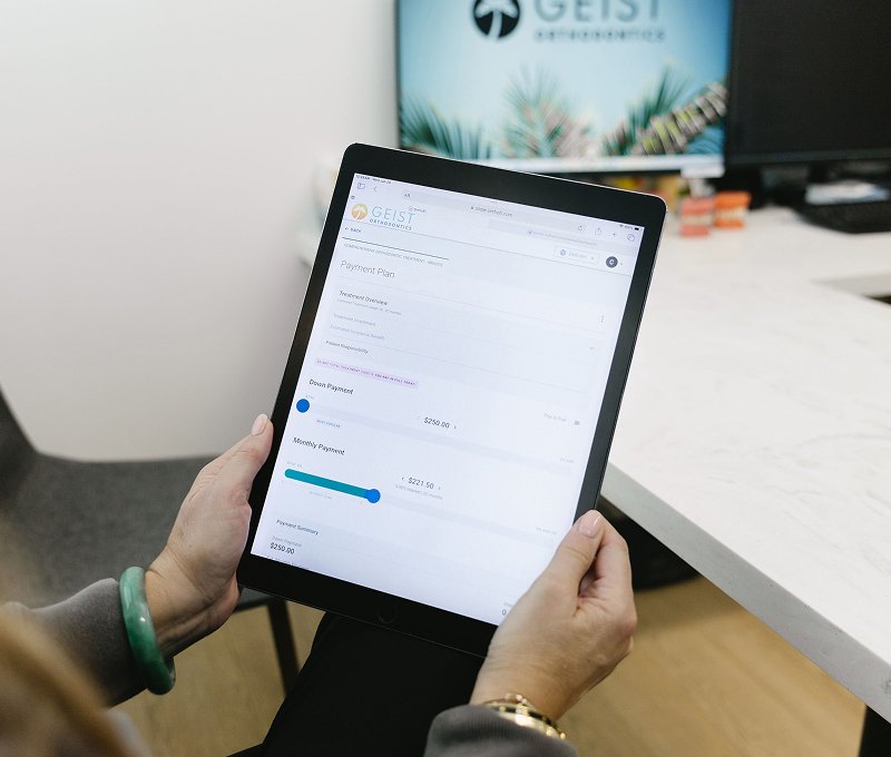 Person holding a tablet displaying a payment plan for orthodontic services at Geist Orthodontics, with a focus on monthly payment options and deposit details, in a modern office setting.